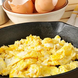 Scrambled eggs in iron skillet next to whisk and broken shells